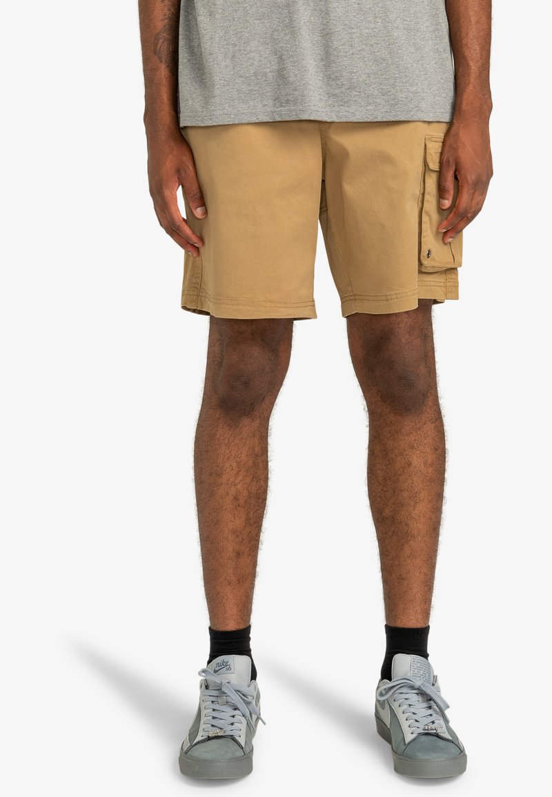 Beige cargo shorts with side pockets, made from cotton. Features straight fit and stitched hem, paired with grey sneakers and black socks.