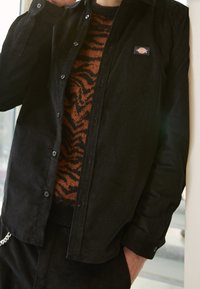 Camicia di velluto a coste nera con bottoni, indossata sopra un maglione a righe arancioni e marroni. Presenta un logo a toppa sulla camicia.