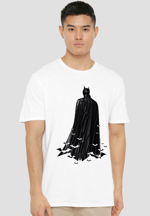 Junger Mann trägt ein weißes T-Shirt mit einer schwarzen Batman-Figur und fliegenden Fledermäusen auf der Vorderseite.
