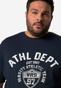 Navy modrá tričko s krátkým rukávem vyrobené z bavlny, s bílým opotřebovaným potiskem s nápisem "ATHL DEPT" a designem štítu s rokem a čísly.