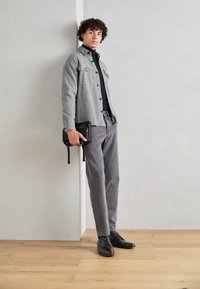 Chemise grise à boutons portée sur un col roulé noir, pantalon gris et chaussures noires. Tient un petit sac noir avec des fermetures éclair et des sangles.