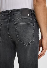 Uomo che indossa jeans in denim grigio scuro con tasche posteriori visibili e una toppa nera con logo sulla vita, camicia bianca parzialmente visibile.