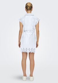 Abito in satin bianco con colletto, maniche corte, vita con cintura e orlo scallopato con motivo floreale intagliato al laser.