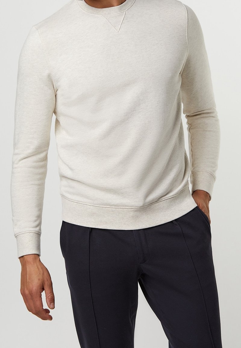 Homme portant un pull à manches longues beige clair et un pantalon bleu marine foncé, une main dans la poche, avec un fond neutre.