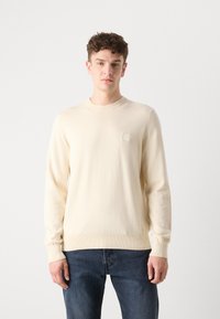Cream-colored knit sweater with a round neckline, ribbed cuffs, and a small branded patch on the chest. Worn with dark blue jeans.