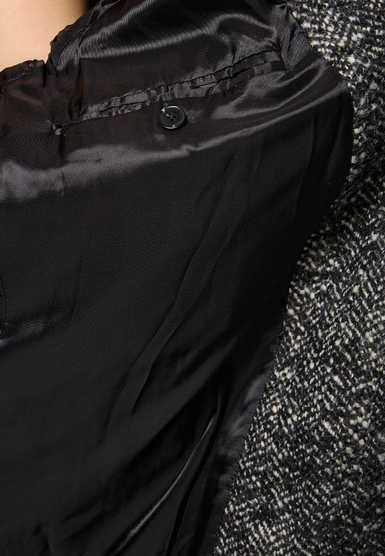 Zwarte satijnen stof met een knoopdetail verschijnt onder een gestructureerd zwart-wit tweed kledingstuk, dat duidelijke contrasten in materialen toont.