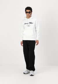 Hvit hettegenser med svart logo, kengurulomme og snøring. Kombinert med svarte bukser og grå/hvite joggesko. Modell med mørke solbriller på.