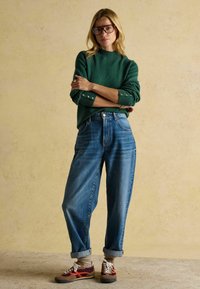 Roheline kudum, mille varrukad on üles keeratud, helesinised kõrge vöökohtadega denimpüksid ja pruunid sneakersid punaste detailidega. Üksik taust.