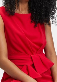 Vestido rojo con un cuerpo ajustado, una cinturilla ancha y un detalle de lazo en la cintura. Hecho de una tela suave y texturizada con sutiles drapeados.