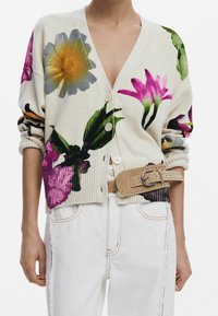 Floral cardigan in cream knit, featuring large purple, yellow, and green flowers. Button closure, ribbed cuffs, and worn with a beige studded belt.