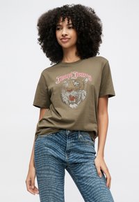 Joven con cabello rizado que lleva una camiseta verde con un gráfico de tigre y texto, y jeans azules con patrones de impresión repetidos de la marca.