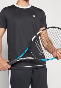 Svart idrottströja med vit krage, med en liten logotyp på bröstet. Modell som håller en tennisracket med blå accent.