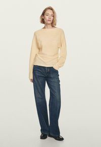 Donna in piedi con una mano in tasca, indossando un maglione beige chiaro, jeans a gamba larga blu scuro e scarpe nere basse.