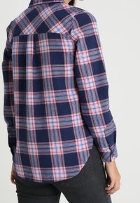 Navy blue and pink plaid flannel shirt with long sleeves, buttoned cuffs, and rounded hem. Features a yoke design at the back.