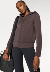 Καφέ φούτερ με φερμουάρ και κουκούλα με ρύθμιση, ριγωτά μανίκια και λεπτομέρεια λογότυπου. Φορεμένο με μαύρες κολάν, κρατώντας μια μαύρη τσάντα γυμναστηρίου.