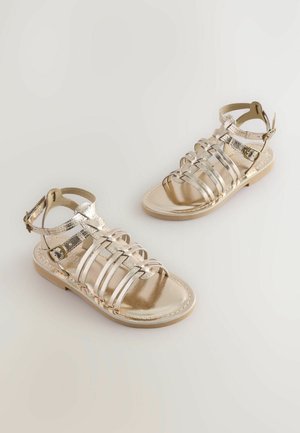 Goldene metallische Sandalen mit flachen Sohlen, die mehrere schmale Riemen und einen verstellbaren Knöchelriemen für Passform und Unterstützung bieten.