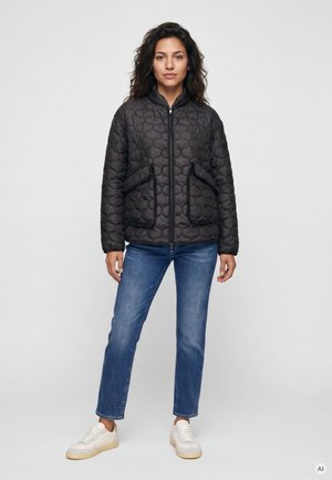 Frau steht und trägt eine schwarze gesteppte Jacke mit Vordertaschen, blaue Jeans und weiße Sneakers vor einem einfarbigen hellen Hintergrund.