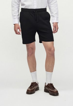 Homme portant un short noir avec cordon, une chemise blanche à manches longues, des chaussettes blanches et des chaussures marron à lacets, debout devant un fond uni.
