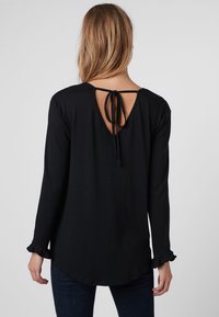 Schwarzes Langarmshirt mit Rundhalsausschnitt und offener Rückenpartie, die mit einem Band gesichert ist. Besitzt geraffte Manschetten und einen lockeren Sitz. Glatte Stoffoberfläche.