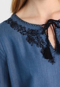Blouse en denim avec un décolleté en V profond et une broderie florale noire. Elle comporte une fermeture à nouer avec des glands, présentant une texture lisse et une couleur bleue.