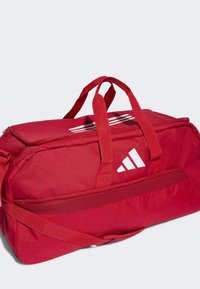 Borsa duffel rossa con manici doppi, tracolla regolabile e tasca in rete, caratterizzata da un logo Adidas bianco e un motivo a righe.