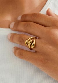 Kuldne sõrmus kolme voolava, tilga kujuga elemendiga. Sile, peegeldav pind ümarate servadega, kantud sõrmel hele kangataustal.