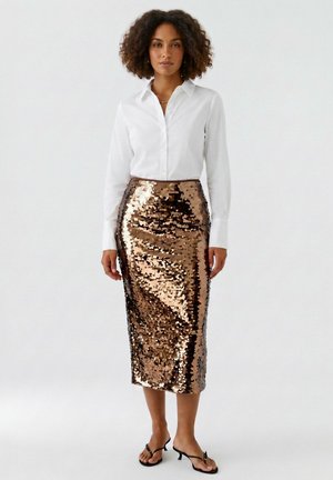 Femme debout portant une chemise blanche à boutons, une jupe midi bronze brillante à sequins, et des sandales à talons ouvertes noires, sur un fond uni.