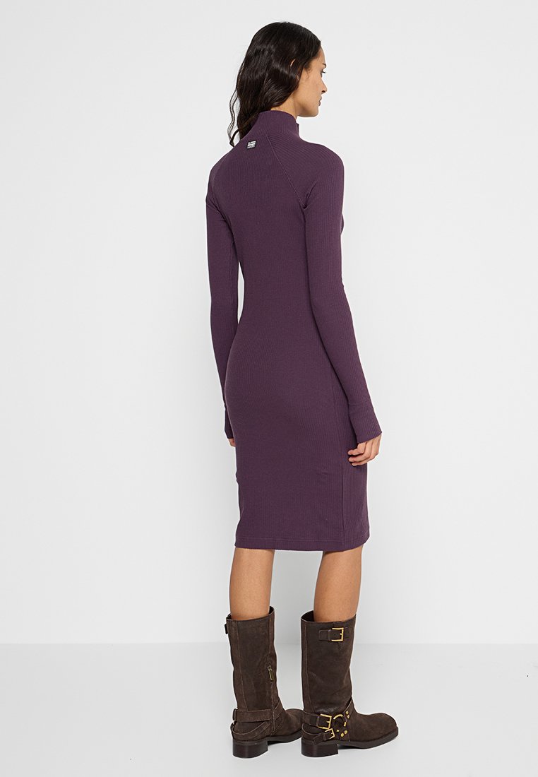 Dopasowana, prążkowana fioletowa sukienka z wysokim kołnierzem i długimi rękawami, noszona z brązowymi botkami do połowy łydki z klamrami i teksturowanym materiałem.