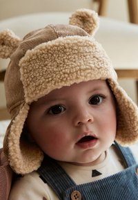 Kapuca iz medvedjega pliša v barve rjave in bež, z puhastimi poudarki na ušesih, teksturiranim flisom in kordurojskim dizajnom. Lepa podrobnost na ušesih in razširjene ušne zaklopke.