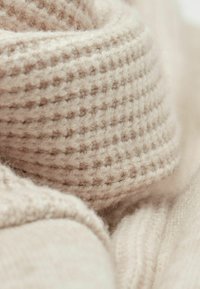 Zbliżenie miękkiej, beżowej dzianiny o teksturowanym, prążkowanym wzorze i widocznymi włóknami, tworzącymi przytulną powierzchnię tekstylną.