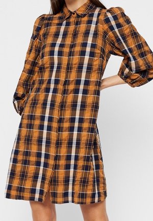 Femme portant une robe à carreaux orange, bleu marine, blanche et beige, longueur genou, avec des manches bouffantes aux trois-quarts et une fermeture boutonnée à l'avant.