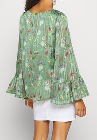 Blusa floreale verde con lunghe maniche a campana, caratterizzata da strisce verticali testurizzate e un orlo arricciato. Tessuto leggero con motivi floreali multicolori.