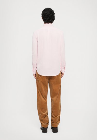 Cămașă roz deschis cu mânecă lungă și hem rotunjit, purtată cu pantaloni de catifea în tonuri de maro, cu broderie florală. Botine negre completează look-ul.
