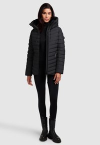 Black puffer jacket with a zip closure, high collar, and hood. Ribbed texture with vertical baffle design, paired with black leggings and boots.