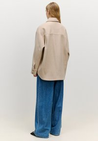 Beige oversized Jacke mit Kragen, rückseitiger Nahtdetails und langen Ärmeln, kombiniert mit lockeren blauen Denim-Hosen. Glatte Textur.