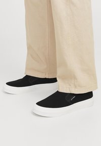 Fekete slip-on sportcipők sima szövetből készült felsőrésszel és fehér gumitalppal, világos bézs bővebb nadrággal párosítva.