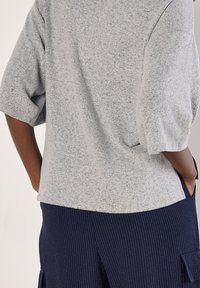 Hellgrauer, strukturierter Pullover mit kurzen Ärmeln, kombiniert mit dunkler, fein gestreifter Hose. Der Pullover hat eine lockere Passform und einen abgerundeten Saum.