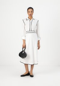 Robe blanche avec des piping noirs, col à boutons, manches longues et taille ajustée. Sac à main noir tenu à la main. Chaussures plates noires.