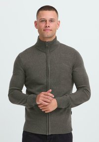 Mann mit kurzen Haaren und Creolen, der einen grauen, ribbeligen Reißverschluss-Pullover trägt und mit gefalteten Händen vor einem schlichten weißen Hintergrund steht.