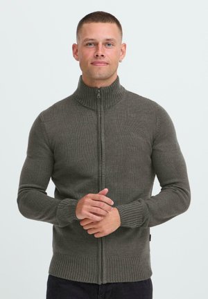 Mann mit kurzen Haaren und Creolen, der einen grauen, ribbeligen Reißverschluss-Pullover trägt und mit gefalteten Händen vor einem schlichten weißen Hintergrund steht.
