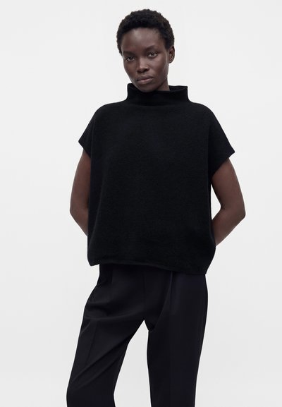 Femme aux cheveux courts portant un pull noir ample sans manches à col roulé et un pantalon noir sur fond blanc uni.