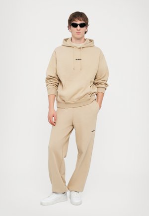 Felpa beige con tasca a marsupio e logo nero, abbinata a pantaloni larghi coordinati, completata da sneakers bianche e occhiali da sole scuri.