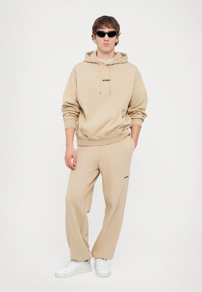 Beige hoodie met kangoeroezak en zwart logo, gecombineerd met bijpassende losse broek, aangevuld met witte sneakers en donkere zonnebril.
