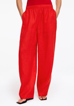 Žena nosi voľné červené ľanové nohavice s bočnými vreckami a čierne sandále s otvorenou špičkou, stojaca na bielom pozadí.