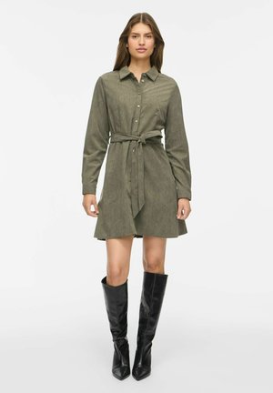 Femme portant une robe-chemise ceinturée à manches longues vert olive et des bottes noires montantes jusqu’aux genoux, debout sur un fond blanc uni.