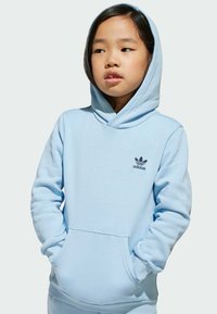 Svijetloplavi dukserica od mekanog materijala, s kenguruvim džepom, velikom kapom i malim crnim Adidas logom na prsima.