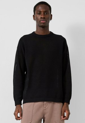 Młody mężczyzna ubrany w czarny dzianinowy sweter z długim rękawem i jasnobrązowe spodnie, stojący na tle gładkiego, jasnoszarego tła.