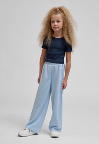 Ung pige med langt krøllet hår iført en sort kortærmet top, lyseblå vidde bukser og hvide sneakers, stående på en grå baggrund.