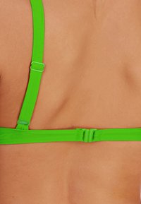 Grüne verstellbare BH-Träger mit einer glatten, glänzenden Textur, ausgestattet mit Kunststoffbeschlägen und einem Schnallenverschluss. Hautfarbe ist im Hintergrund sichtbar.