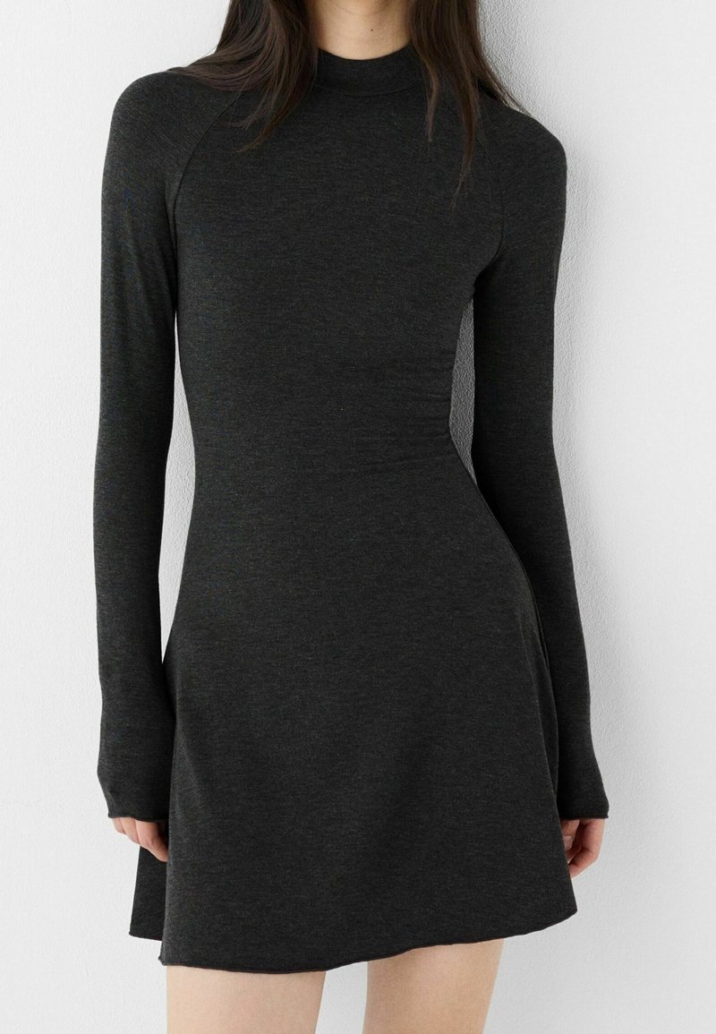 Femme portant une robe en maille gris foncé, cintrée, à manches longues et col montant, debout contre un mur blanc uni.